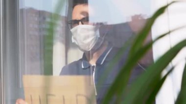 30 yıllık adam mavi tişört giyiyor, koruyucu tıbbi tek kullanımlık yüz maskesi takıyor, pencereden dışarı bakıyor. Hüzünlü izole edilmiş hasta elinde karton tabela, elinde de yazı var. Kişisel izolasyon kavramı. 