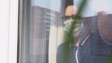 Hasta adam tek kullanımlık beyaz yüz maskesi takıyor, pencereye geliyor, cama yaslanıyor, dışarı bakıyor, üzerinde el yazısıyla yazılmış bir yazı olan kartonu tutuyor. Bir salgın sırasında evde yalnızlık.