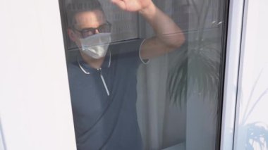 Üzgün yalnız esmer adam koruyucu tıbbi yüz maskesi takıyor, koyu renk tişört camdan dışarı bakıyor, cama doğru itiyor. Coronavirus covid-19 virüsü karantinaya almış. Grip bulaşmış. Kapalı