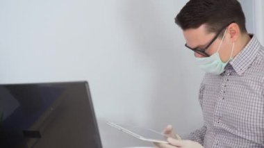 Genç bir erkek yönetici gri gömlek giyiyor, siyah gözlük takıyor, koruyucu yüz maskesi takıyor, dizüstü bilgisayar kullanıyor, video konuşmasında konuşuyor, tablette grafiğini gösteriyor, insanlara öğretiyor. Evde güvenli bir uzaktan kumanda. Kilitle..