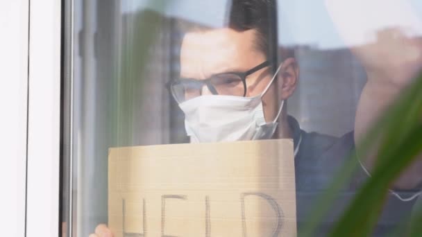 暗いTシャツ 医療面マスクを身に着けている普及した若い白人男性が窓の外に立って 碑文ヘルプで署名を保持し ガラスの上に傾きます Guy Quarantine Coronavirus Covid Virus Threat — ストック動画
