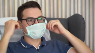 Koltuğunda oturan yakışıklı bir esmer adam. Tek kullanımlık tıbbi turkuaz yüz maskesi takıyor. Çizgili arka planda siyah gözlükleri var. Beyaz adam karantina döneminde evde kalıyor..