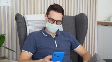 Mutlu genç adam tıbbi maske takıyor, siyah gözlükler takıyor, kablosuz kulaklıklar takıyor, rahat bir koltukta oturuyor. Memnun adam telefonu elinde tutuyor. Erkek rahatla, müzik dinle. Yüzünde mutluluk.