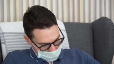 Genç hasta esmer adam ARVI virüsünden kalma tek kullanımlık tıbbi maske takıyor, siyah klasik gözlükler takıyor, kulaklarına kablosuz kulaklık takıyor, müzik dinliyor, yakın mesafeden sandalyeye oturuyor.