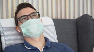 Kısa kahverengi saçlı beyaz adam tıbbi yüz maskesi takıyor, siyah gözlükler takıyor, koltukta oturuyor, kulaklarında kablosuz kulaklıkla müzik dinliyor. Mutlu adam gülümser, başını sallar evde müzikal vuruşlara