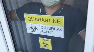 Mavi tişörtlü hasta izole bir adam pencere camına yaslanmış ve üzerinde sarı uyarı yazısı yazılı Quarantin ALERT. Adam maske takıyor, öksürüyor, hastane sağlık odasından dışarı bakıyor. Yüzü yok