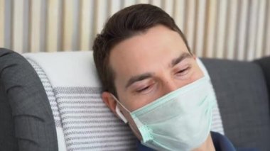 Beyaz kulaklıklı, gri koltuklu hasta bir gencin portresi. Adam koruyucu yüz maskesi takıyor, izolasyon ve salgın sırasında evde kalıyor. Sonbahar, akut solunum viral enfeksiyon zamanı.