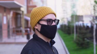 Güneş gözlüklü genç adam, siyah gazlı bez koruyucu yüz maskesi, sarı örgülü şapka sokakta bulanık şehir arka planına karşı duruyor. Adam dikkatlice yana bakıyor, derin derin nefes alıyor, gözlüklerden duman çıkıyor..