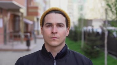 Cazibeli, ciddi, traşlı, koruyucu siyah maske takan, üzerinde kırmızı çizgi yazılı Covid-19, sonbahar şapkası olan genç bir adam. Bulanık sokaktaki hasta erkek portresi yakın plan..