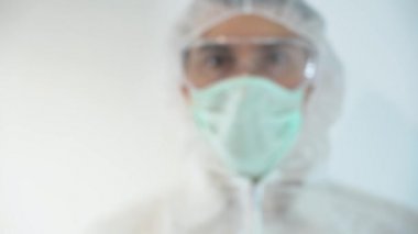 Bulanık bilim adamının portresi, koruyucu elbise giyen sağlık görevlisi, yüz maskesi, gözlük, beyaz arka planda eldivenler. Doktor virolog elini kaldırıyor, kan testi tüpü gösteriyor. Aşı, hastalık tedavisi.
