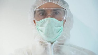 Kendine güvenen genç adam koruyucu giysi giyiyor, tek kullanımlık yüz maskesi, gözlük, kafaya kaput, mavi lastik eldiven, cam test tüpü gösteriyor..