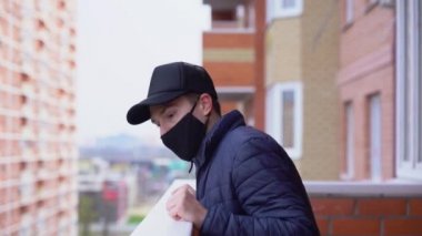 Çekici genç erkek balkonda duruyor, aşağı bakıyor, koruyucu maskeyi çıkarıyor, aç gözlülükle temiz hava soluyor. İyileşme kavramı, virüsten kurtulma. Sonbahar Sonbaharı.