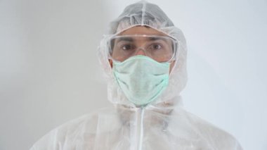 Korkak doktor portresi koruyucu giysi, eldiven, turkuaz yüz maskesi, gözlük, beyaz arka planda başlık giyer. Adam elinde tutuyor, dikkatlice kan koronası virüslü test tüpüne bakıyor..