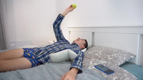 ある幸せな若いハンサムな怠惰な白人のブルネットの男性は 青の再生シャツ パンティー リラックスして ベッドの上に枕の上に横たわって 天井を見て 自宅で黄色のテニスボールを投げます — ストック動画