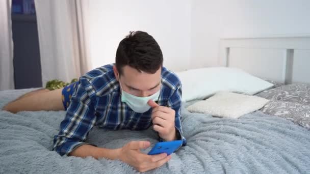 短い茶色の髪を持つ1人だけで退屈大人の男は スマートフォンを使用して 灰色のベッドに横たわって 保護顔マスク パンツ 青のシャツを着用します ウイルスのパンデミックによる隔離期間中の自己分離 — ストック動画