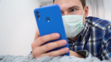Kapalı portre genç esmer adam, dinlenirken, yatakta uzanır, akıllı telefon kullanır. Mutlu adam mavi ekose gömlek giyiyor, tıbbi PPE yüz maskesi takıyor, telefonu tutuyor, internetten ilginç videolar izliyor. Seçici odak.