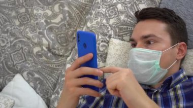 Hasta, yakışıklı, beyaz, esmer adam mavi ekose gömlek giyiyor, turkuaz, tek kullanımlık koruyucu yüz maskesi takıyor, elinde telefonla yatakta yatıyor. Üst Manzara. Uzayı kopyala Erkek evde kalır..