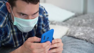 Hasta, yakışıklı esmer adam turkuaz, tek kullanımlık tıbbi koruyucu yüz maskesi, mavi ekose gömlek giyiyor, yatağa uzanıyor, akıllı telefon kullanıyor, internette sörf yapıyor. Uzayı kopyala İzolasyon konsepti. Salgın Günleri.