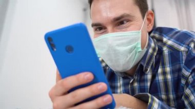 Kahverengi gözlü mutlu esmer bir adamın yakın plan portresi tek kullanımlık tıbbi yüz maskesi takıyor, mavi kareli flanel gömlek giyiyor ve akıllı telefonuyla eğlendirici içeriği izliyor.