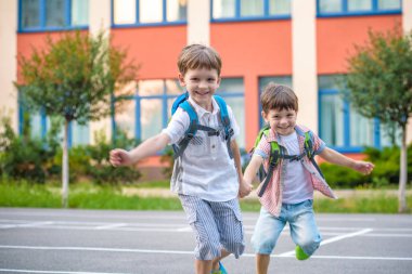 Young students, two sibling brothers, going to school. They hold hands. Children behind shoulders have satchels. Warm day in an early autumn. Back to school. Happy smiling kids. clipart