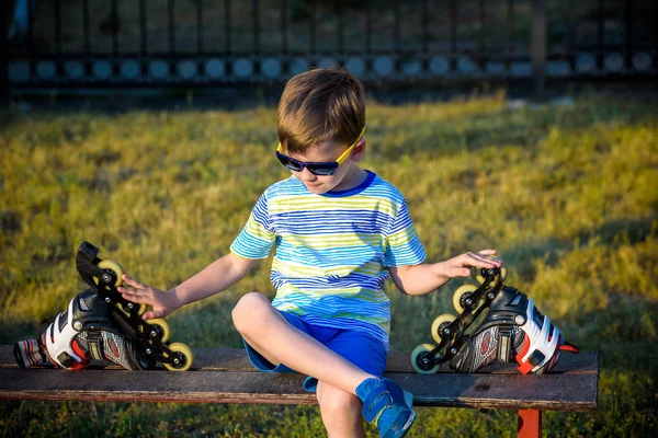 Little handsome boy in roller-blades sits on bench in summer park. Happy smile sport kid in sunglasses wear safety helmet protection. Active leisure summer holidays time with family.