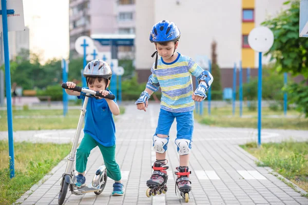 Two kid boy on roller skates and his sibling brother on scooter wrapped in park. Children wearing protection pads for safe roller skating ride. Active outdoor sport for kids.