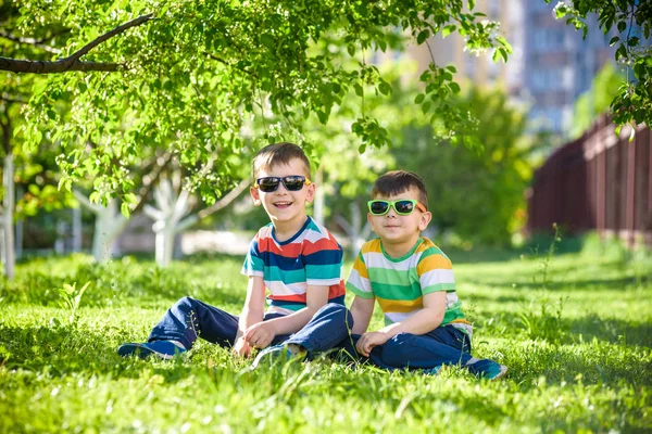 Mutlu yaz tatili. İki çocuk bir yaz Park yeşil çim üzerinde. Kardeş kardeşi çocuklar çiçek açan ağaçlar arka plan üzerinde güneş gözlüğü takıyor Park'ta oturuyor. Çocuklar mutlu ve gülümseyerek dostluk kavramı.