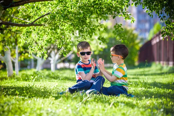 Mutlu yaz tatili. İki çocuk bir yaz Park yeşil çim üzerinde. Kardeş kardeşi çocuklar çiçek açan ağaçlar arka plan üzerinde güneş gözlüğü takıyor Park'ta oturuyor. Çocuklar mutlu ve gülümseyerek dostluk kavramı.