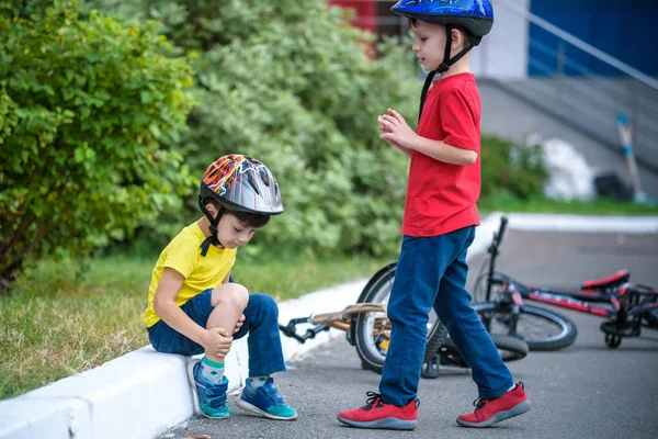 小男孩从自行车上摔下来 不快乐的男孩坐在柏油路上看着他的膝盖 他的朋友帮他站起来 又骑行了 — 图库照片