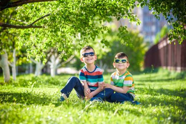 Mutlu yaz tatili. İki çocuk bir yaz Park yeşil çim üzerinde. Kardeş kardeşi çocuklar çiçek açan ağaçlar arka plan üzerinde güneş gözlüğü takıyor Park'ta oturuyor. Çocuklar mutlu ve gülümseyerek dostluk kavramı.