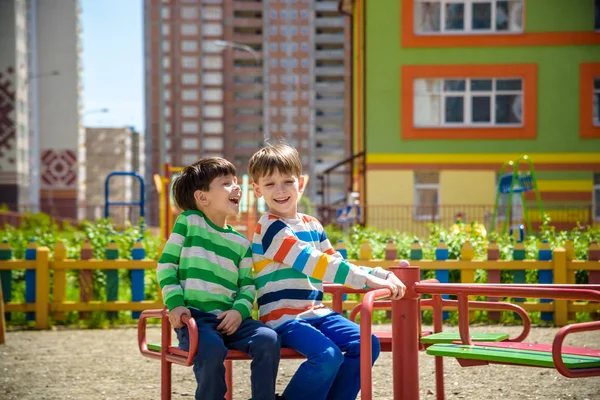 Dos chicos alegres en camisetas juegan en un gran carrusel de hierro en el patio de recreo en el verano o el día cálido de primavera en el jardín de infantes. Los niños se divierten mucho. Concepto de amistad —  Fotos de Stock