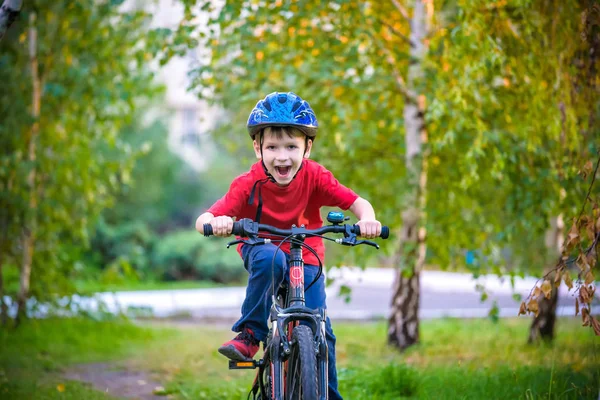 Um menino branco pequeno da criança em um capacete protetor em sua cabeça senta-se em uma bicicleta das crianças. A criança em uma bicicleta vermelha de duas rodas olha para a frente. Um sorriso astuto no rosto da criança — Fotografia de Stock