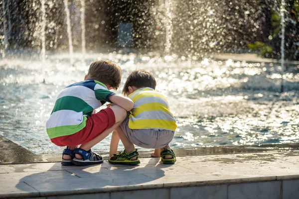 Lindo niño y hermanos mayores, jugando en una fuente de chorro con agua salpicando alrededor, verano — Foto de Stock