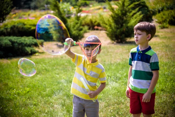 Chico soplando burbujas de jabón mientras un niño emocionado disfruta de las burbujas. Feliz adolescente y su hermano en un parque disfrutando haciendo burbujas de jabón. concepto de amistad infancia feliz — Foto de Stock