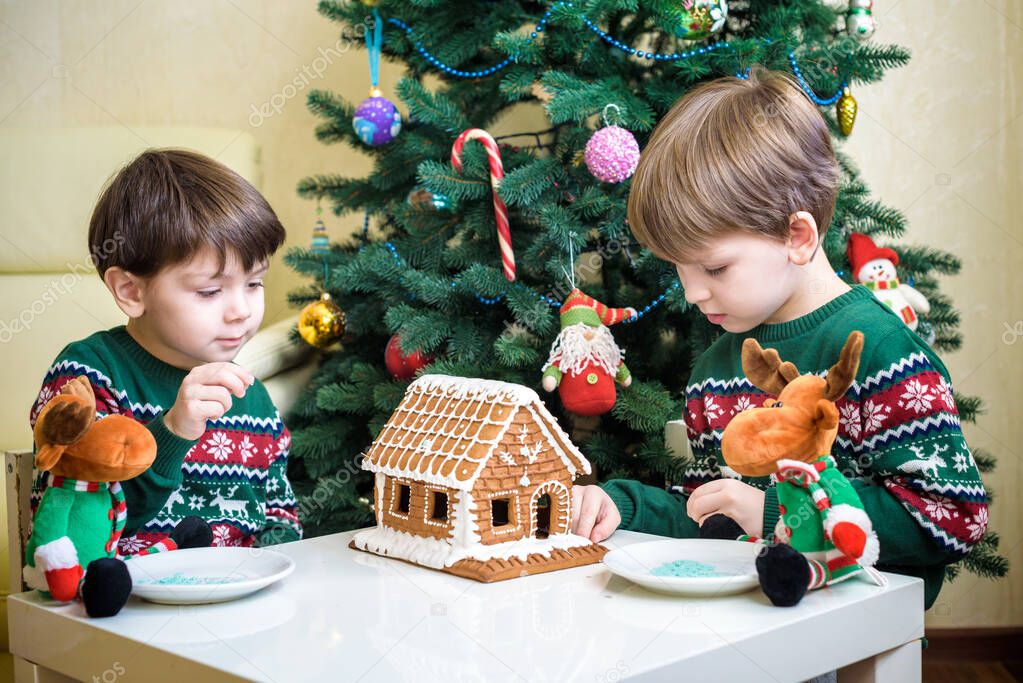 Two sweet boys, brothers, making gingerbread cookies house, decorating at home in front of the Christmas tree, child playing and enjoying, Christmas concept