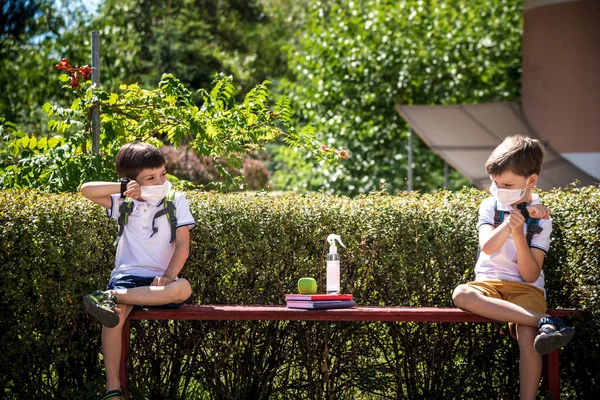 Coronavirus school reopening concept: a school boys sit on bench in front of a school wearing a face mask. Communicating trough smart watch. Covid-19, health, education, safety, and back to school.