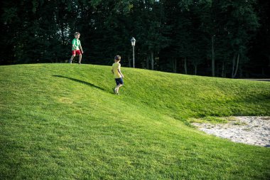 Golf sahasında koşan iki çocuk arka plandaki yeşil çimlerde ve ormandaki tepelerde koşuyor..