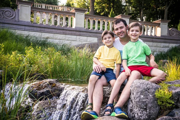 Baba ve iki çocuk, küçük oğlan ve şelale geçmişi olan açık hava portresi. Kristal deresinin yanındaki büyük kayanın üzerinde..