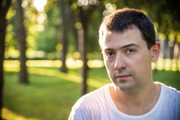 Açık havada, beyaz tişörtlü, doğa parkının arka planında güzel, ciddi bir adamın portresini çek. Moda modeli.