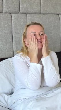 Kadın yüzüne ve ellerine krem sürerken mutlu mesut yatağında oturur, kendine dikkat eder beyaz yataklı Avrupalı orta yaşlı kadın gülüşü. Yüksek kalite 4k görüntü