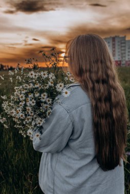 Bir kız tarlada yürüyor, ve bir buket küçük beyaz papatya çiçeğiyle poz veriyor, arka planda yeşil bir tarla ve turuncu bir gün batımı, uzun sarı saçlı bir kız