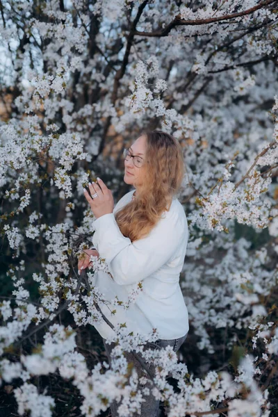 Fotoğrafta beyaz çiçeklerle açan bir ağacın yanında poz veren bir kız görüyorsunuz.