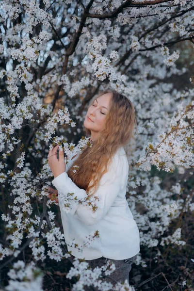 Fotoğrafta beyaz çiçeklerle açan bir ağacın yanında poz veren bir kız görüyorsunuz.