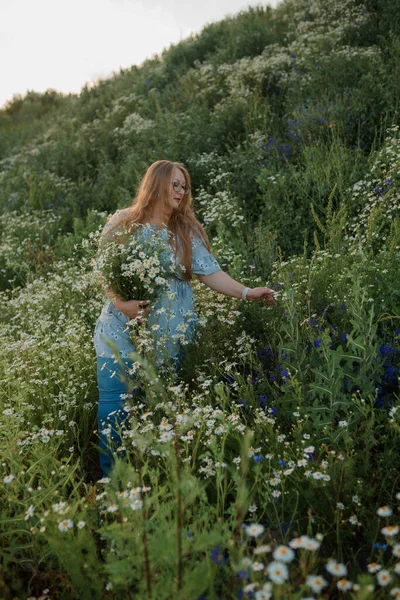 Fotoğrafta uzun sarı saçlı bir kız tarlada yürüyor ve küçük beyaz bir papatya buketi topluyor.