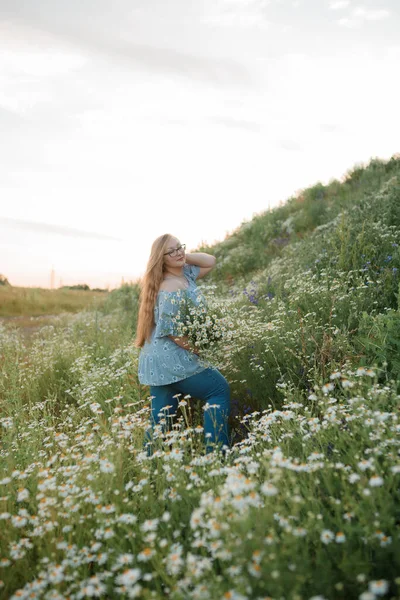 Fotoğrafta uzun sarı saçlı bir kız tarlada yürüyor ve küçük beyaz bir papatya buketi topluyor.