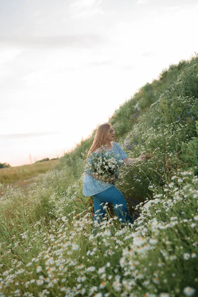 Fotoğrafta uzun sarı saçlı bir kız tarlada yürüyor ve küçük beyaz bir papatya buketi topluyor.