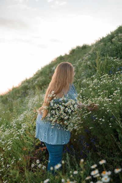 Fotoğrafta uzun sarı saçlı bir kız tarlada yürüyor ve küçük beyaz bir papatya buketi topluyor.