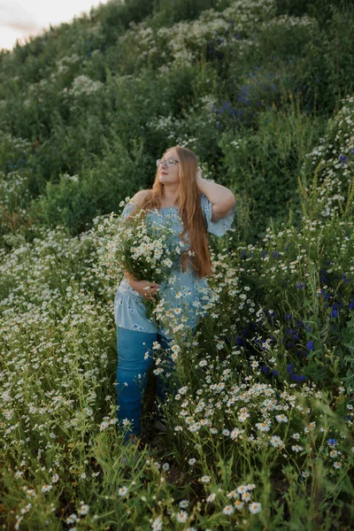 Fotoğrafta uzun sarı saçlı bir kız tarlada yürüyor ve küçük beyaz bir papatya buketi topluyor.