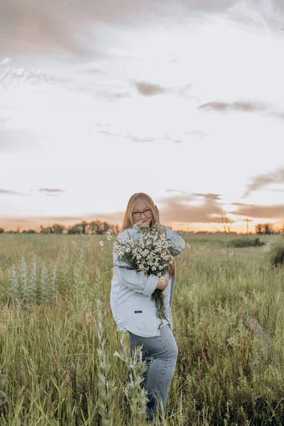 Bir kız tarlada yürüyor, ve bir buket küçük beyaz papatya çiçeğiyle poz veriyor, arka planda yeşil bir tarla ve turuncu bir gün batımı, uzun sarı saçlı bir kız