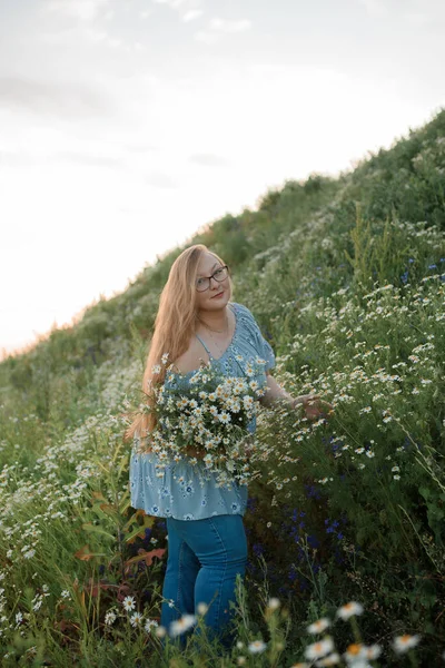 Fotoğrafta uzun sarı saçlı bir kız tarlada yürüyor ve küçük beyaz bir papatya buketi topluyor.